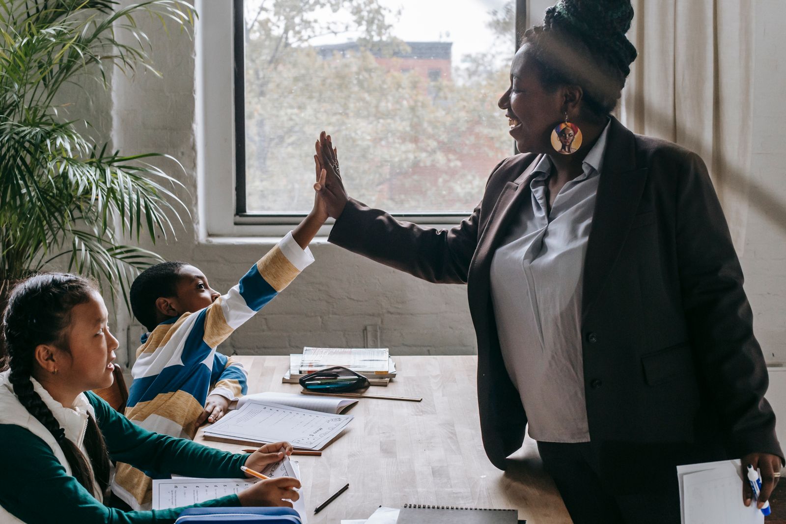 A teacher sharing a high-five with a smiling pupil at a desk in a bright classroom while classmates look on.