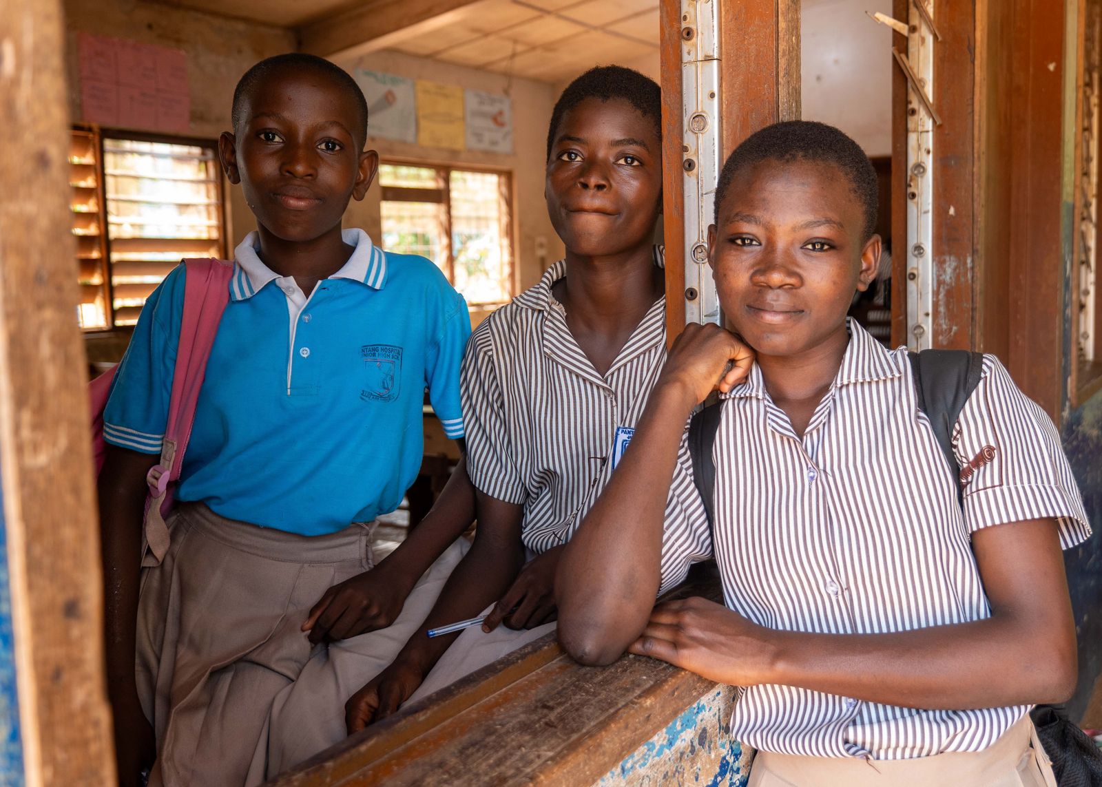 Three teenage secondary-school students smiling together inside their classroom.