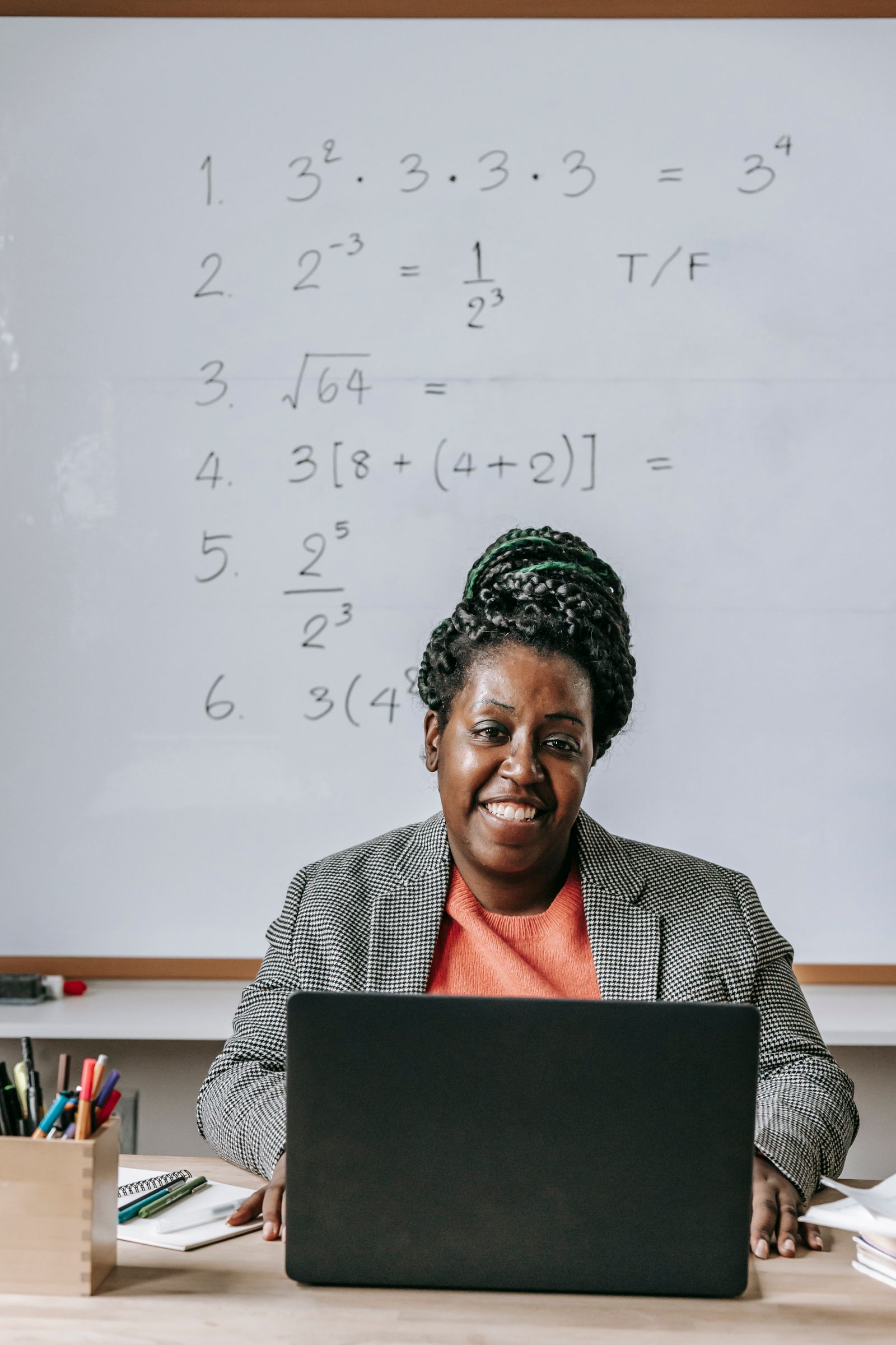 A cheerful educator seated at a classroom desk working on a laptop and smiling at the camera.